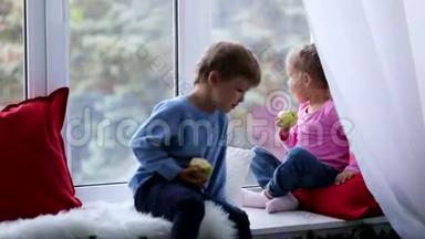 兄妹俩坐在窗台上，吃着苹果，看着窗外。